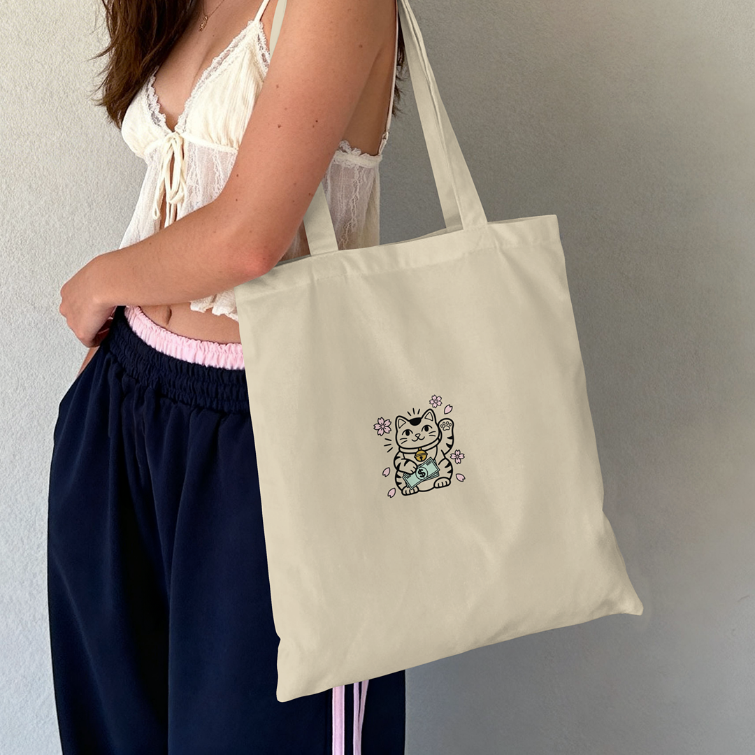 Lifestyle photo of a woman carrying the Hustle Hard Lucky Cat embroidered canvas tote bag.