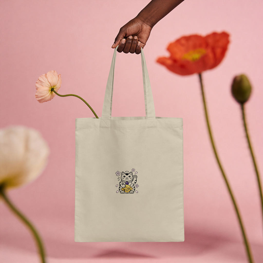 Hand holding the Wealth Magnet Lucky Cat embroidered canvas tote bag against a floral pink background.